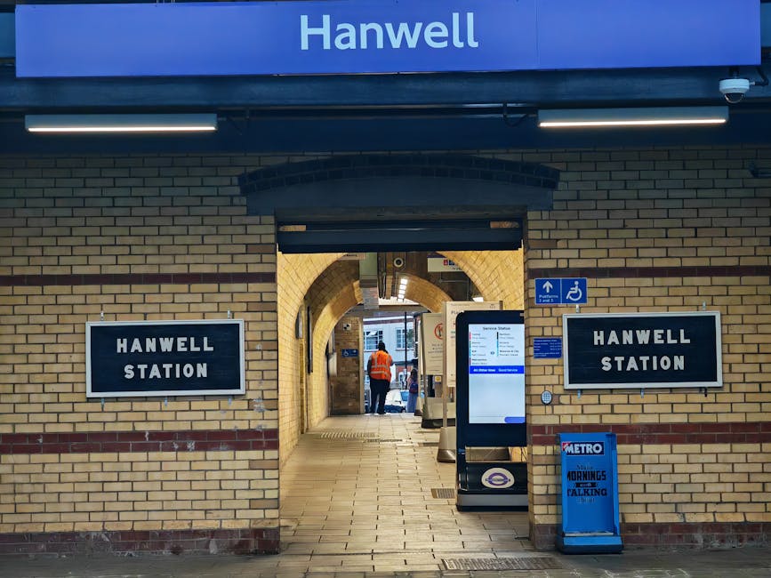 Interior view of Hanwell Station entrance with a brick archway leading to the platform area. The station features beige and red brickwork on the walls, with two black and white signs reading 'Hanwell Station' positioned on either side of the entrance. In the foreground, there is a digital ticket machine and a blue Metro sign indicating the morning and talking service. Inside the station, a person wearing an orange high-visibility vest is seen walking along the tiled pathway, which shows a clear area for loading and unloading during home relocations or furniture transport. The environment is well-lit with fluorescent lighting fixtures, and the scene captures a moment during a house removal or luggage transfer process facilitated by professional removals, such as Man and Van Abbey Wood, assisting with packing and moving logistics near this transport hub.