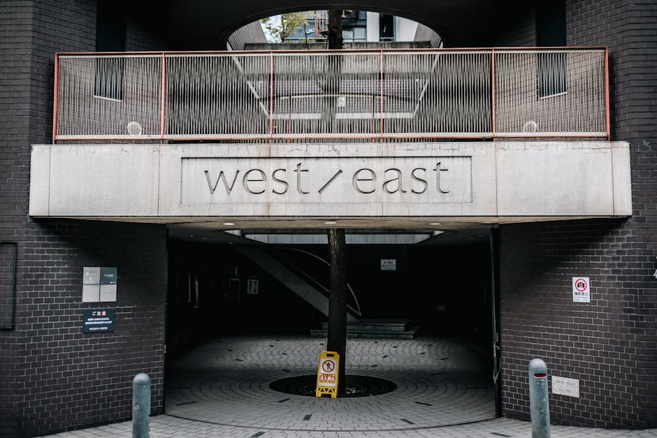 A multi-storey residential or commercial building with a large concrete overhang bearing the engraved text 'west/east' on its front facade. The structure features dark brick walls and a balcony with a metal railing above the entrance. The entrance itself is an open archway leading into a shaded parking or underpass area, with a staircase visible within. In front of the archway, there is a round landscaped area with a tree growing from the pavement, surrounded by cobblestone-patterned paving. A yellow and black safety sign is placed around the tree, and there are bollards positioned on either side of the entrance to manage traffic or pedestrian flow. The overall scene appears to be part of a contemporary urban setting, relevant to building access and logistics for home relocation or furniture transport, as managed by companies like Man and Van Abbey Wood.
