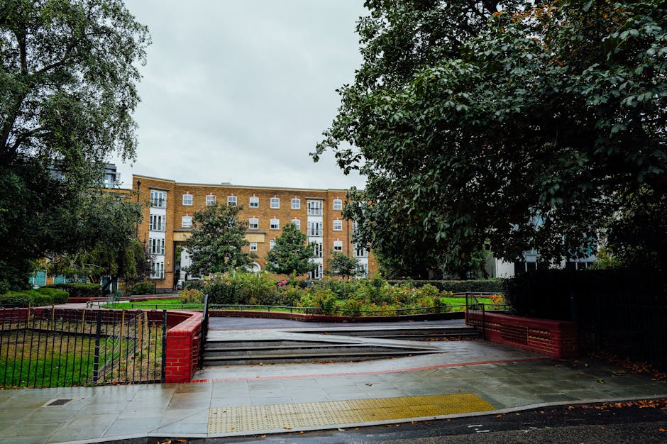 The image shows a residential courtyard with a paved pathway leading to a set of metal stairs, which are partially blocked by a red brick wall and black metal railing. To the right, there is a large tree with dense green foliage, and on the left, another tree with hanging branches. In the background, a multi-story brick apartment building with white-framed windows and balconies is visible, surrounded by smaller trees and shrubbery. The sky above is overcast, creating diffused lighting across the scene. The courtyard appears to be part of an urban residential area, possibly where furniture or boxes might be loaded and unloaded during house removals or home relocation activities. Man and Van Abbey Wood offers services in this area, addressing access challenges for basement flats and similar properties, supporting smooth furniture transport and packing during moving processes.