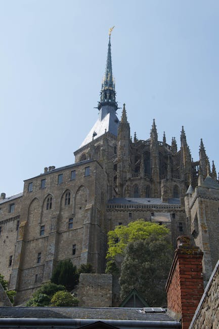 A tall, historic stone building with Gothic architectural features, including a steep, pointed spire topped with a gold weather vane, situated on a hillside. The structure has multiple levels with arched windows, buttresses, and decorative stonework. Surrounding the building are green trees and shrubs, with a brick chimney visible at the bottom right corner. The overall environment appears to be a clear day with blue sky, and the image captures the building from a low angle, emphasizing its height. This scene relates to house removals or relocation services, as it portrays a significant, well-known structure often associated with historic or residential settings, relevant to the services provided by Man and Van Abbey Wood.