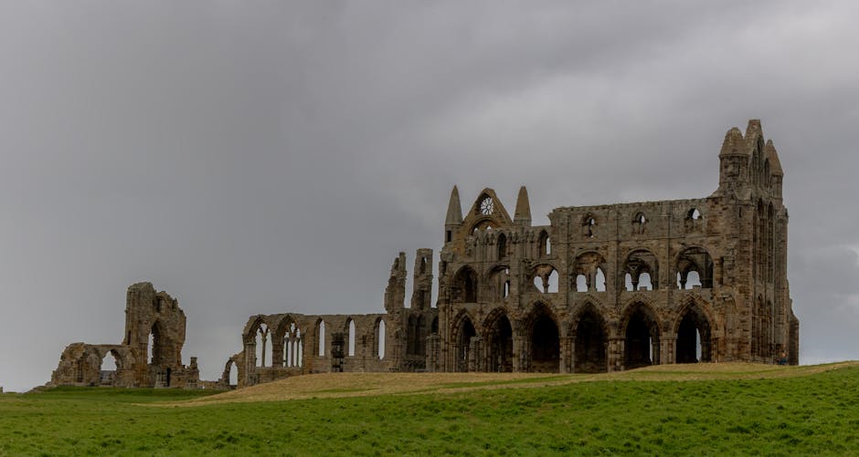A large, historic stone ruin located atop a gently sloping grassy hill, featuring multiple arched windows and doorways, with some sections partially collapsed and others standing tall. The structure includes Gothic architectural elements, such as pointed arches and tall, narrow windows. Dark, cloudy skies loom overhead, casting diffuse lighting over the scene. This historic building is viewed from a distance, with the foreground consisting of green grass and the background of overcast skies, highlighting its weathered stone walls and intricate architectural details. Man and Van Abbey Wood may use such images to illustrate the importance of careful packing and transport when handling valuable or delicate heritage or antique items during house removals, especially for properties near historic sites.