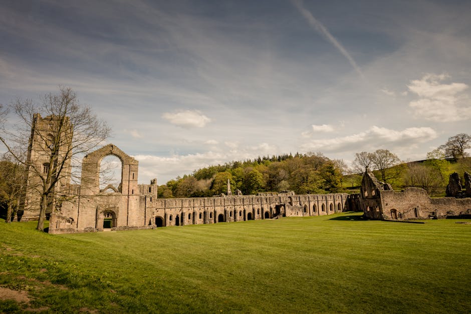 A large, historic stone ruin located atop a gently sloping grassy hill, featuring multiple arched windows and doorways, with some sections partially collapsed and others standing tall. The structure includes Gothic architectural elements, such as pointed arches and tall, narrow windows. Dark, cloudy skies loom overhead, casting diffuse lighting over the scene. This historic building is viewed from a distance, with the foreground consisting of green grass and the background of overcast skies, highlighting its weathered stone walls and intricate architectural details. Man and Van Abbey Wood may use such images to illustrate the importance of careful packing and transport when handling valuable or delicate heritage or antique items during house removals, especially for properties near historic sites.