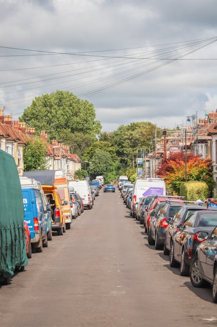 A residential street scene during daylight hours showing parked cars lining both sides of the narrow road, including various models of sedans, hatchbacks, and vans. The kerbside features a row of houses with front gardens, some decorated with bushes and small trees, and a few houses displaying brick and stucco facades. Overhead, a series of power lines stretch across the cloudy sky. In the distance, a blue van with the branding of Man and Van Abbey Wood is partially visible, positioned near the pavement, suggesting a home relocation or moving process underway. The environment appears calm, with minimal pedestrian activity, and the street is free of obstructions, ideal for careful furniture transport during house removals. The scene subtly highlights typical urban moving logistics, including vehicle positioning and street accessibility in preparation for a house relocation, in line with services such as those offered by Man and Van Abbey Wood.