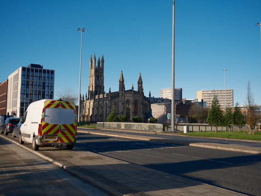 A white van with high-visibility red and yellow chevron markings on the rear is parked on the roadside in front of a historic church with tall, ornate spires. The van is positioned near the pavement, adjacent to a busy urban street with multiple lanes and parked cars, some of which are visible along the side. Modern office buildings and residential apartments surround the church, with a few trees and grassy areas enhancing the scene. Tall streetlights are evenly spaced along the road, and the sky is clear and blue, indicating daytime. The scene depicts typical city relocation logistics, with the van possibly being used for house removals or furniture transport, exemplifying the logistics involved in packing and moving processes, as seen by the presence of commercial vehicles associated with professional removals services like those offered by Man and Van Abbey Wood.