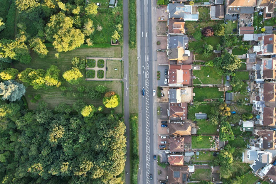 Aerial view of a residential area in Abbey Wood showing a row of detached and semi-detached houses with small back gardens, some with sheds and patio furniture, situated alongside a main road with par