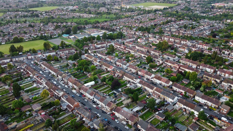 Aerial view of a residential area in Abbey Wood showing a row of detached and semi-detached houses with small back gardens, some with sheds and patio furniture, situated alongside a main road with par
