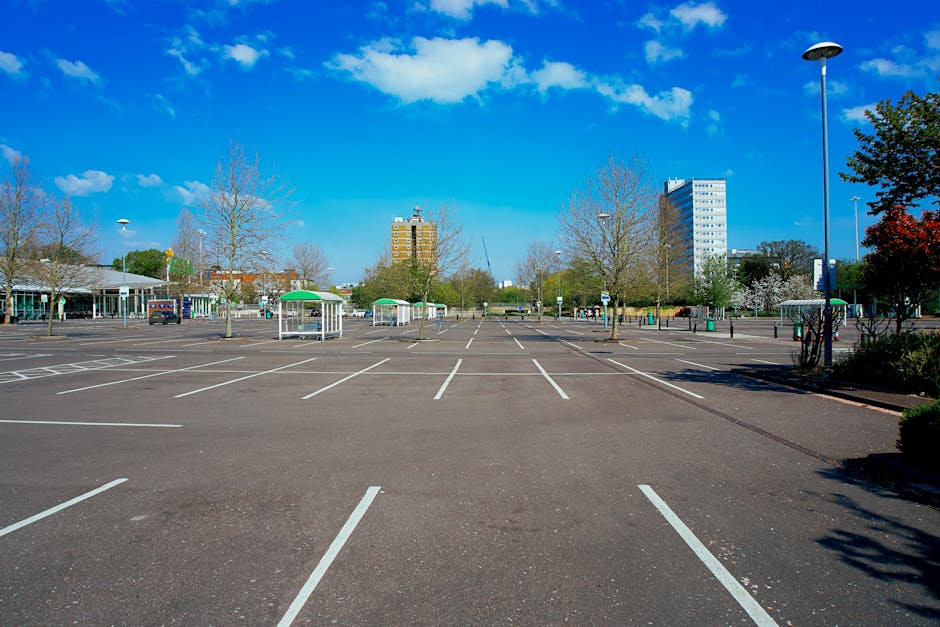 An empty parking lot situated outdoors on a clear day with blue skies and a few scattered clouds. The lot features numerous designated parking spaces marked with white lines, some of which are occupied by a few vehicles parked near the edges. Several small green roofed shelters are positioned within the lot, and a tall streetlamp stands on the right side of the image. Surrounding the lot, there are leafless trees and some shrubs, indicating it might be early spring or late autumn. In the background, multi-storey buildings can be seen, including a taller white or light-colored structure, suggesting an urban or suburban environment. The scene is well-lit, with natural sunlight casting soft shadows. This setting is typical for a public parking area used during home relocation or furniture transport processes, as managed by companies like Man and Van Abbey Wood, situated within the context of house removals and moving services in Greenwich.