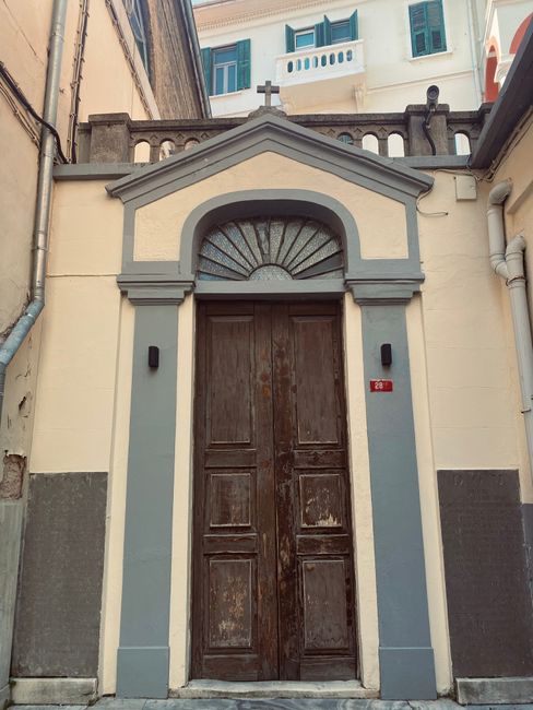 A close-up view of the front entrance of a building featuring a pair of tall, weathered wooden doors with visible signs of wear and peeling paint, set within an arched stone frame. The archway is topped with a semi-circular window made of metal bars arranged in a radial pattern, with a decorative molding above it. Flanking the doors are two black doorbell buttons or intercoms mounted on the wall. The building's facade showcases a combination of cream and grey paint with grey accents outlining the door frame and supporting columns, contrasting with the otherwise smooth plaster surface. Above, part of an upper-floor balcony with a balustrade and windows is visible, suggesting this building may be part of a multi-story structure in an urban setting. The scene is lit naturally, emphasizing the textures of the wood and plaster, and the proximity to other buildings indicates a typical residential or historic area. As part of a home relocation or moving process, this entrance might be used as an access point for furniture transport, with the building's architectural features providing context for logistical planning, especially in tight city spaces. Man and Van Abbey Wood specializes in removals, including careful handling of furniture and packing materials for such locations.