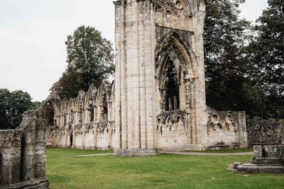 A photograph of the ancient ruins of Lesnes Abbey Woods, featuring a weathered stone wall with Gothic-style arched windows and decorative stone carvings. In the foreground, there is a neatly maintained grassy area and a part of an old stone pillar. Large trees with green foliage surround the ruins, framing the partially collapsed stone structure against an overcast sky. The scene captures the historical architecture of the abbey remains, with visible details of the stonework and natural surroundings. This setting illustrates the kind of scenic environment where a home relocation or furniture transport service might operate, with Man and Van Abbey Wood specialising in removals and moving logistics, often dealing with packing, loading, and transporting items through such historic and outdoor environments.