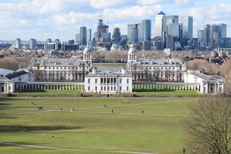 A wide view of a large, white historic building with two prominent towers and rows of colonnades extending across the front, situated behind a vast, well-maintained green lawn with several people walking or sitting. In the background, there is a modern city skyline with numerous tall skyscrapers, some under construction, against a partly cloudy sky. The scene appears to be during daytime with natural lighting. The image showcases a contrast between the classical architecture of the historic house and the contemporary urban environment behind it. The setting suggests a location where house removals or relocation services, such as those offered by Man and Van Abbey Wood, could take place, with moving activities possibly involving careful packing and transportation of furniture and belongings from the historic site to an urban area.
