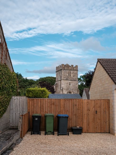 Image showing a small gravel driveway enclosed by a wooden fence and stone walls, with four waste bins in black, green, blue, and black, positioned on the gravel surface. Behind the fence, a historic stone tower with battlements and small windows is visible against a partly cloudy sky. To the right, part of a brick house with a pitched roof is seen, indicating a residential property in Abbey Wood. The scene suggests preparations for a house removal or home relocation process involving loading or waste disposal, with the presence of waste bins as part of the moving logistics managed by Man and Van Abbey Wood, consistent with local regulations for waste collection during removals.