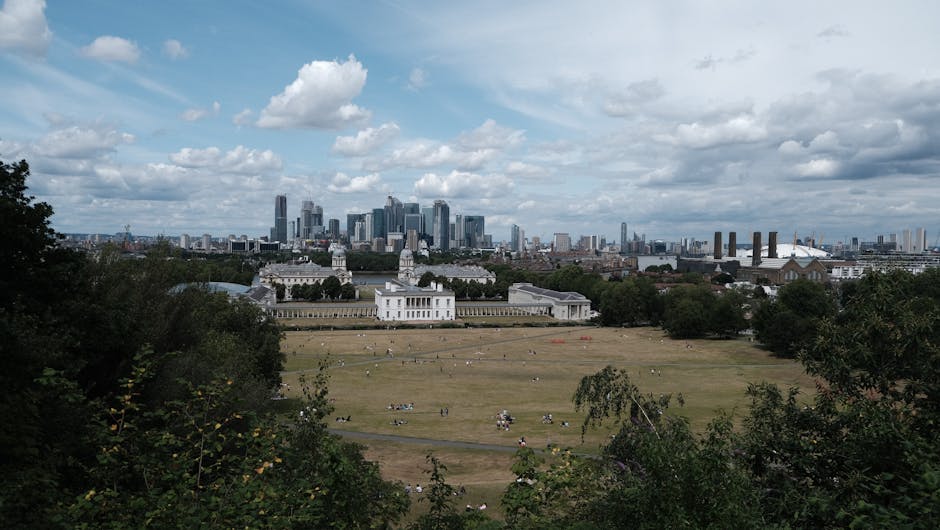 A wide view of a large, white historic building with two prominent towers and rows of colonnades extending across the front, situated behind a vast, well-maintained green lawn with several people walking or sitting. In the background, there is a modern city skyline with numerous tall skyscrapers, some under construction, against a partly cloudy sky. The scene appears to be during daytime with natural lighting. The image showcases a contrast between the classical architecture of the historic house and the contemporary urban environment behind it. The setting suggests a location where house removals or relocation services, such as those offered by Man and Van Abbey Wood, could take place, with moving activities possibly involving careful packing and transportation of furniture and belongings from the historic site to an urban area.