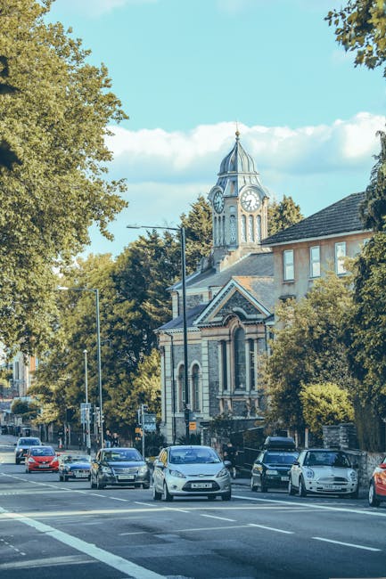 Aerial view of a town centre in Abbey Wood, showing a variety of commercial buildings, shops, and residential houses surrounded by green trees and streets. A curved road with multiple lanes runs throu